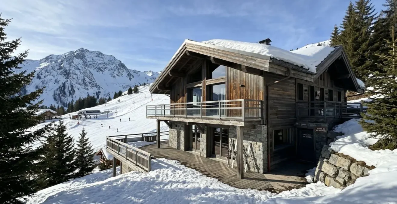 Façade d'un chalet contemporain en bois et pierre avec les pistes de ski enneigées de Courchevel visibles en arrière-plan sous un ciel bleu d'hiver