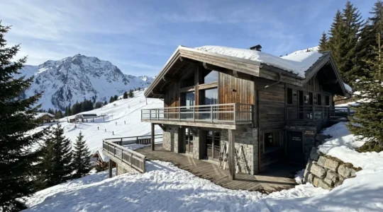 Façade d'un chalet contemporain en bois et pierre avec les pistes de ski enneigées de Courchevel visibles en arrière-plan sous un ciel bleu d'hiver